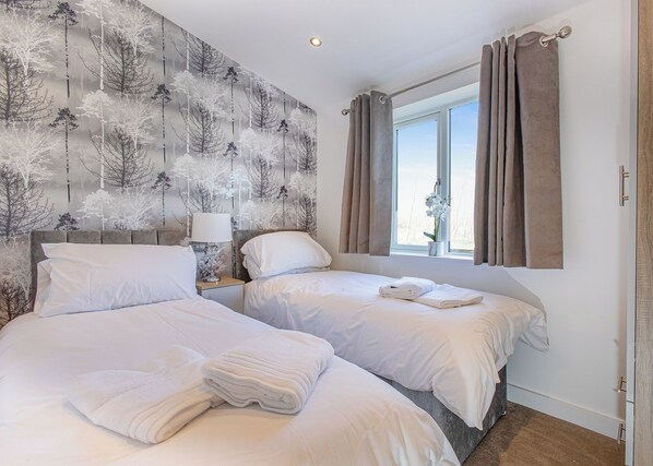Hotellihuone, jossa on kaksi sänkyä, yöpöytä lampulla, ikkuna verhoilla ja seinä, jossa on puukuvioprintti.