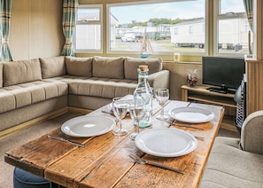A cozy living room with a sectional sofa, a wooden dining table set for two, a television, and a view of mobile homes outside.