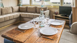 A cozy living room with a sectional sofa, a wooden dining table set for two, a television, and a view of mobile homes outside. - Large 12ft-wide caravan with one double with en suite W. (Cardigan)