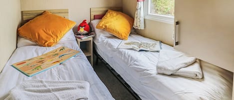 Une chambre avec deux lits, chacun doté de draps blancs et d’oreillers jaunes, une petite table de chevet avec un livre et un objet rouge, et une fenêtre avec des rideaux.