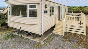 A mobile home with a ramp leading to a gated entrance, a small porch, and a view of the surrounding landscape. - Large 12ft-wide caravan with one double with en suite W. (Cardigan)