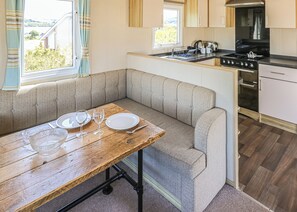 A compact kitchen with a dining area, including a bench, a table with dishes and glasses, and a stove. - Large 12ft-wide caravan with one double with en suite W. (Cardigan)