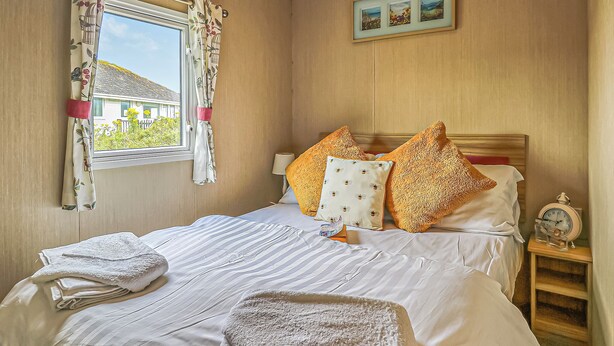 A neatly made bed with pillows, a bedside table with a clock, and a window with curtains. - Large 12ft-wide caravan with one double with en suite W. (Cardigan)