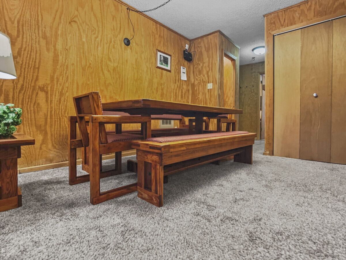 Una habitación con un banco y sillas de madera, suelo alfombrado y paredes de madera, además de una puerta.