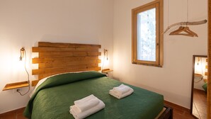 A room with a wooden headboard, a green bedspread, two neatly folded white towels, a window with a wooden frame, and a wooden hanger on the wall.