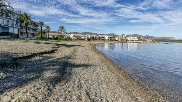 Une plage avec un hôtel en arrière-plan, un ciel dégagé et des montagnes à l’horizon.