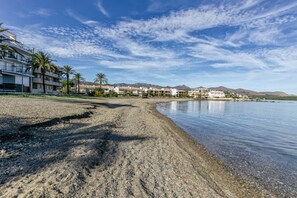 A beach with a hotel in the background, clear skies, and mountains in the distance.