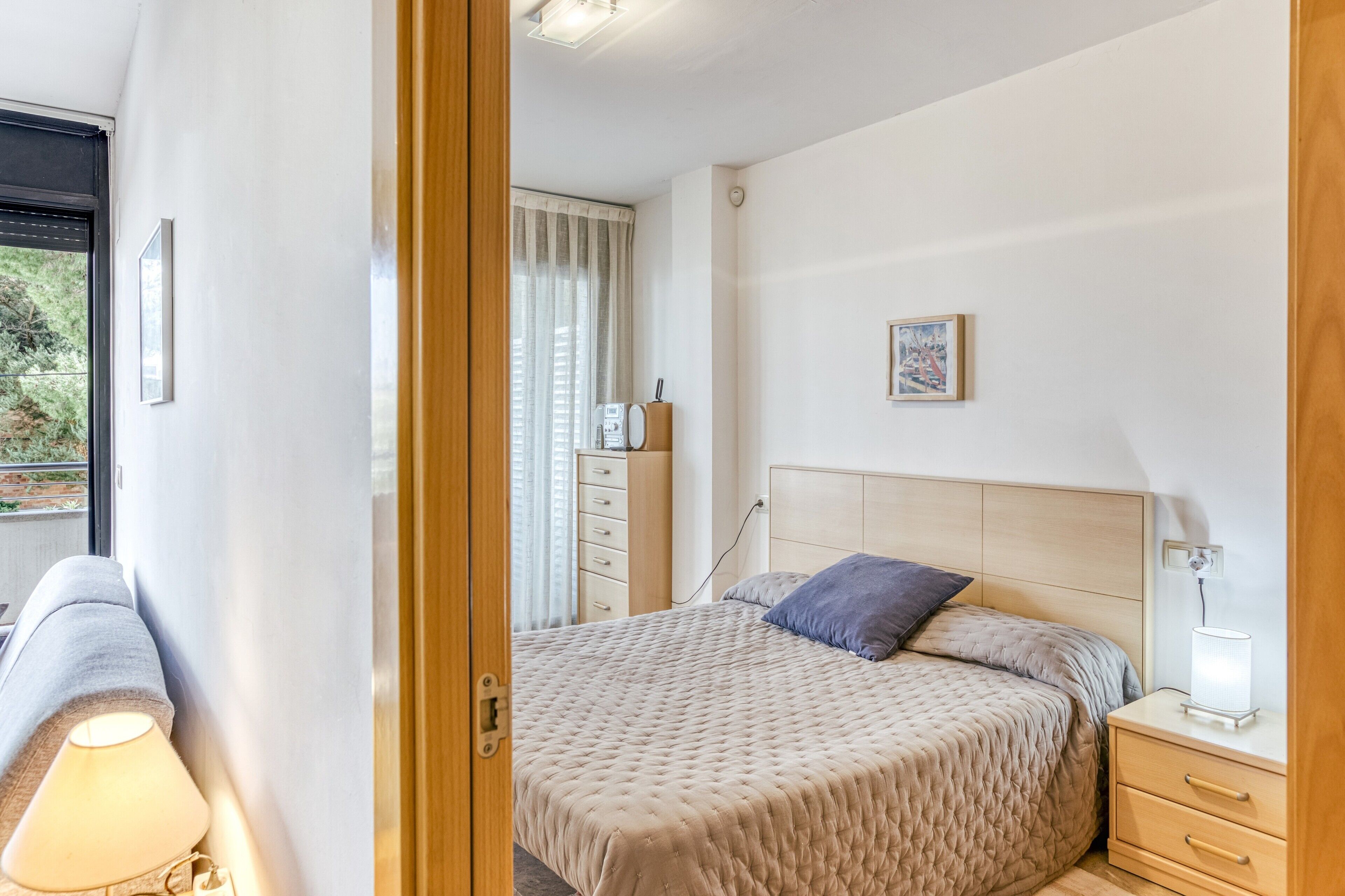 Un dormitorio con cama, mesita de noche, lámpara y una ventana con cortinas.