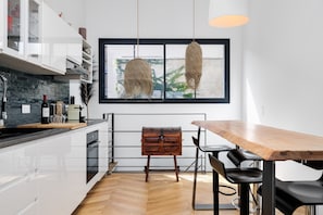 Une cuisine moderne avec des meubles blancs, une table à manger en bois et une grande fenêtre agrémentée de luminaires suspendus.