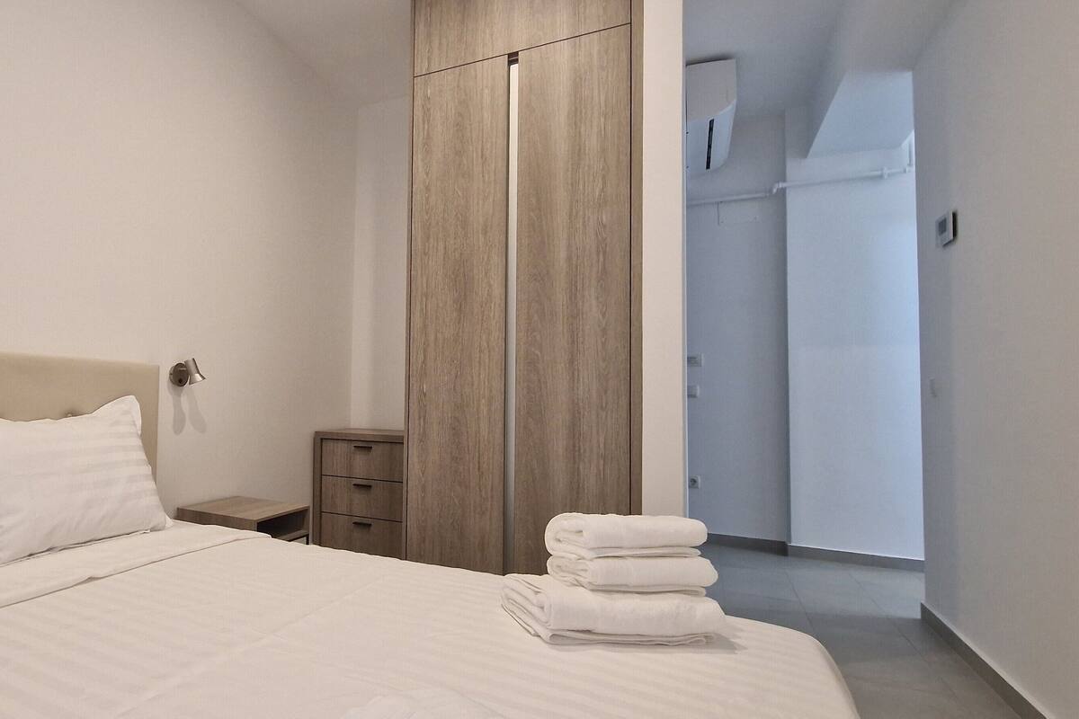 Quarto de hotel moderno com uma cama grande, um guarda-roupa de madeira, uma mesinha de cabeceira e uma cama bem arrumada com toalhas dobradas.