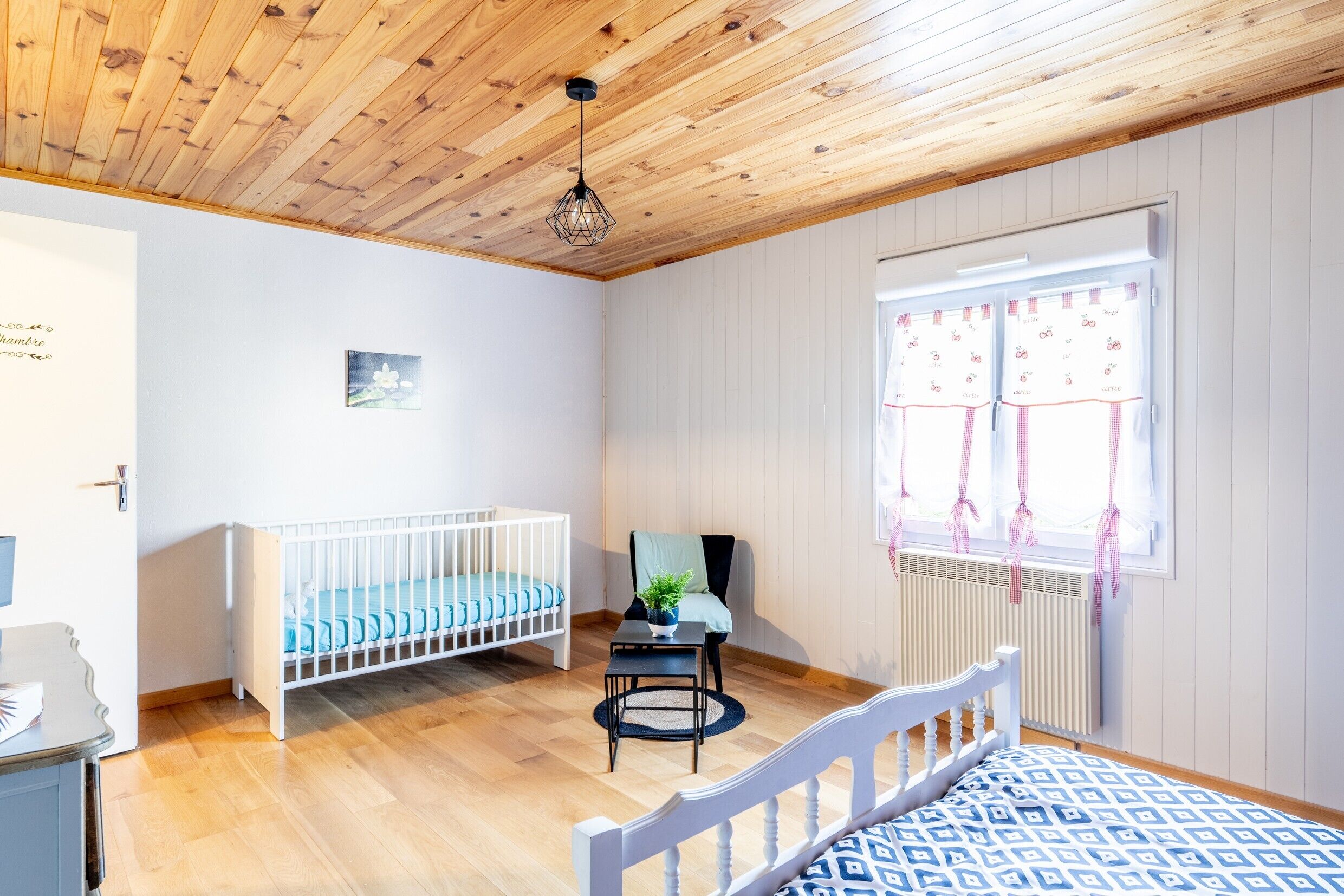 Una habitación con una cuna, una cama, una silla, una mesa y un ventanal con cortinas.