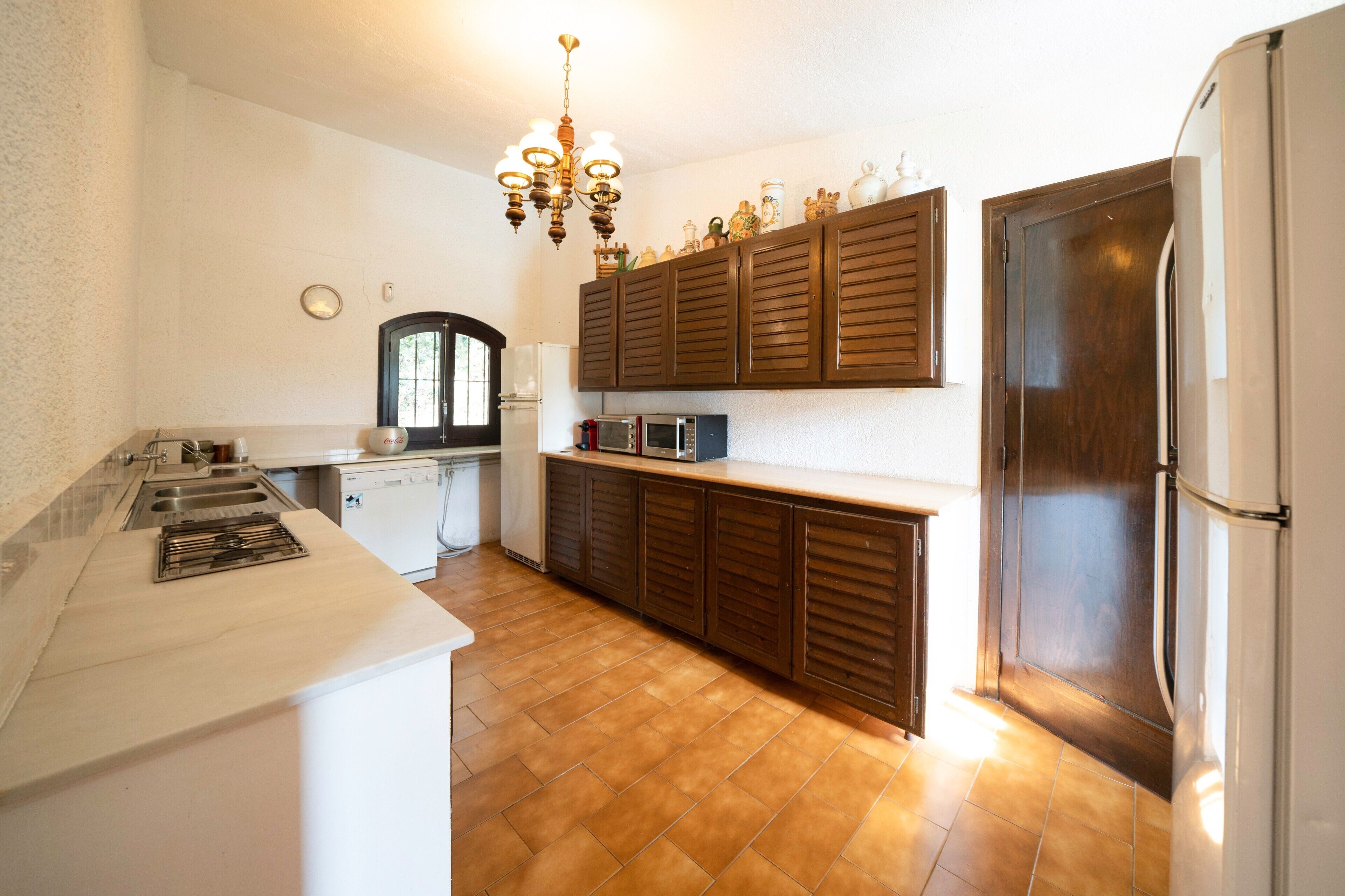 Eine Küche mit Mikrowelle, Backofen und Kühlschrank. Oberhalb der Arbeitsfläche befindet sich ein Holzschrank, an der Decke hängt eine Chandelier.