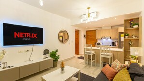 A modern living room with a flat-screen TV displaying the Netflix logo, a dining area with a white table and chairs, and a kitchen in the background.
