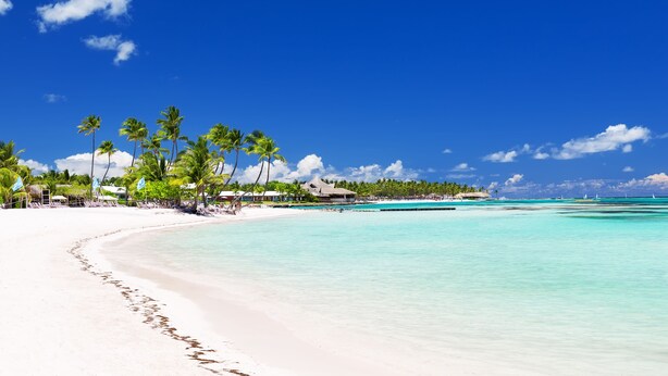 A beach with white sand, clear turquoise water, and palm trees. There are buildings with thatched roofs in the background.