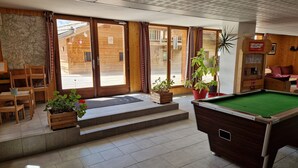 Ein Billardtisch in einem Raum mit Holzboden, einer Tür, die nach draußen führt, und einigen Kübelpflanzen.