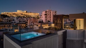 Una terrassa amb una banyera d'hidromassatge tancada amb vidre, vista a l'Acròpolis antiga i edificis moderns al fons.
