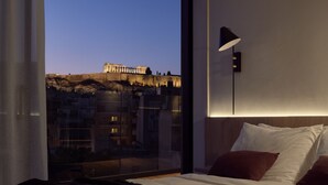 Un dormitori amb una finestra gran, un llit amb roba de llit blanca i marró, una làmpada de nit moderna i vista a un edifici històric.