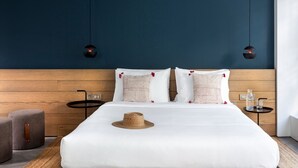 A modern hotel room with a wooden headboard, two bedside tables, a bed with white bedding, a straw hat on the bed, and two pendant lights.