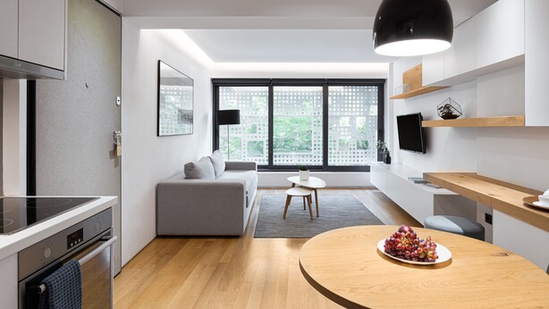 Een moderne keuken met een inbouwoven, een eethoek met een ronde houten tafel en een grijze bank.