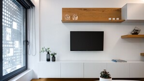 Een moderne woonkamer met een groot raam, een flatscreen televisie aan de wand, houten planken en een ronde witte tafel met een potplant.