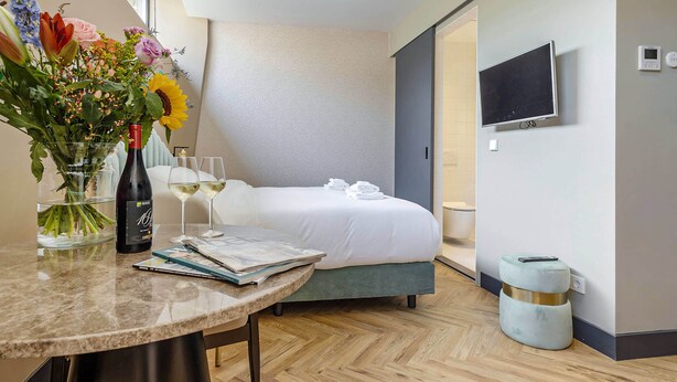 Habitación de hotel con cama, televisión, un florero con flores, botella de vino y una mesa con revistas.
