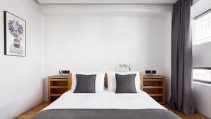 A modern bedroom with a large bed, two bedside tables, a nightstand, a framed artwork, and a window with curtains.