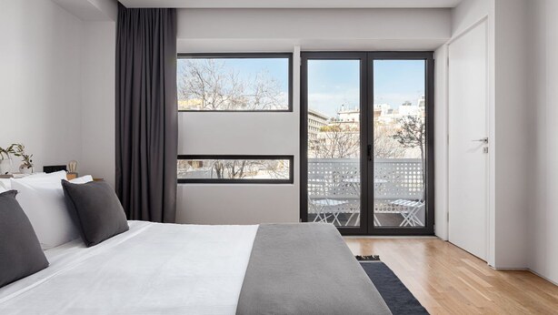 A modern bedroom with a large bed, a view of the city through a sliding glass door, and a balcony with outdoor furniture.