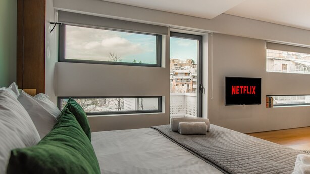Et moderne soveværelse med en stor seng, udsigt over byen og et fjernsyn, der viser "NETFLIX".