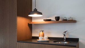 Et moderne køkken med et træskab, en hængende sort lampe, kaffemaskine, to kopper, en vask og en hylde med skåle og krukker.