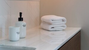 Et badeværelse med bordplade i hvid marmor, en vask, et spejl og en stak håndklæder. Der er to flasker shampoo og balsam på bordet.
