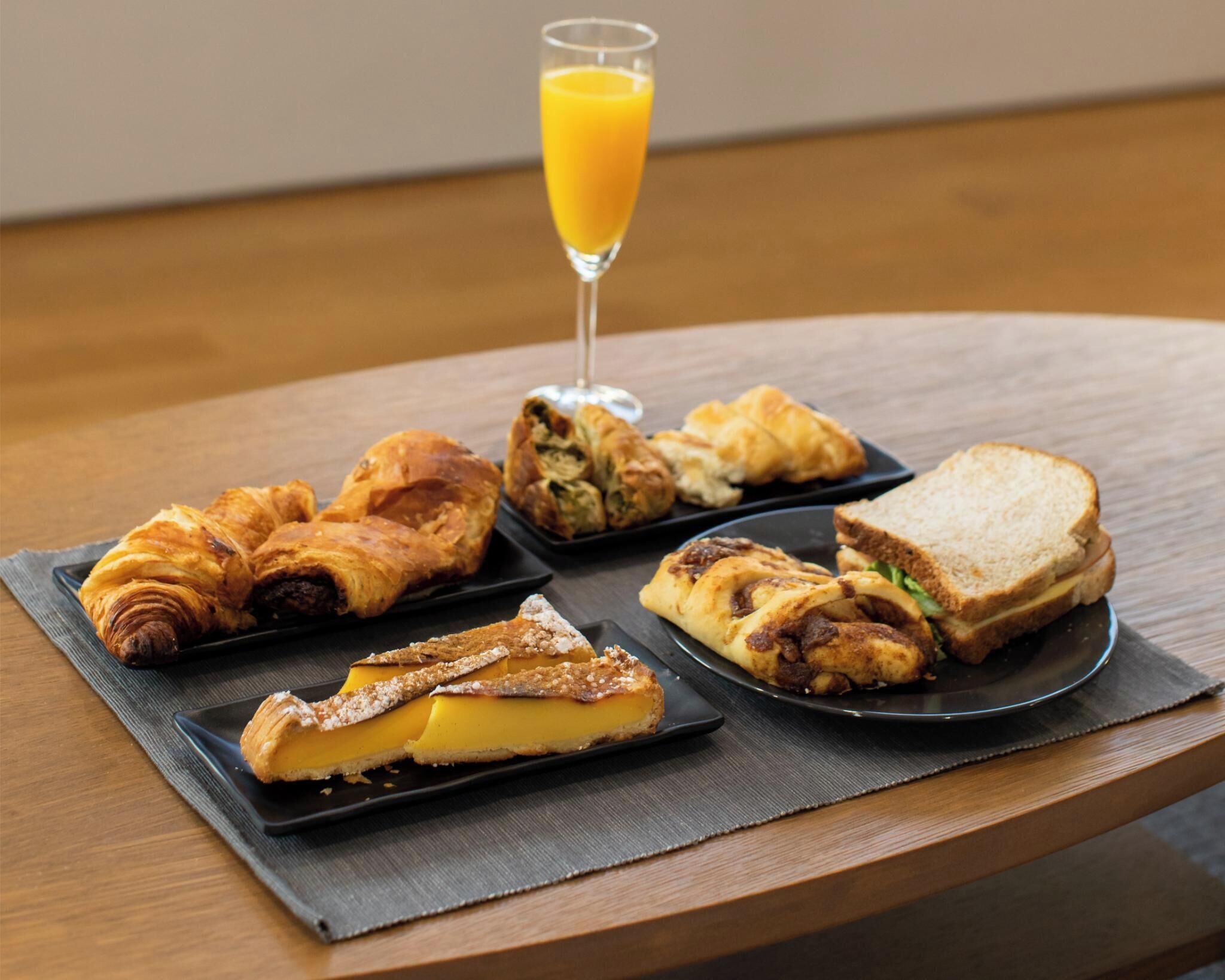 A breakfast setting with croissants, a cheese tart, a sandwich, and a glass of orange juice on a wooden table.