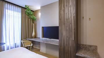 A hotel room with a bed, a desk with a flat-screen TV, a chair, and a large plant.