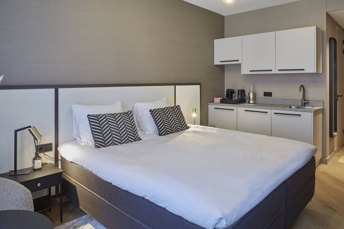 A modern hotel room with a large bed, a bedside table with a lamp, a small desk, and a kitchenette area with cabinets and a sink.