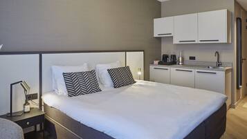 Una habitación de hotel moderna con una cama grande, una mesita de noche con lámpara, un escritorio pequeño y una zona de kitchenette con armarios y un lavamanos.