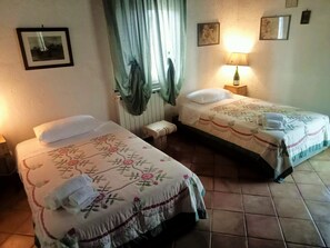 Two single beds with floral bedspreads, a small bedside table, a lamp, and a window with curtains. - Cozy villa with views of the river, the village of Saturnia and Montemerano. (Saturnia)