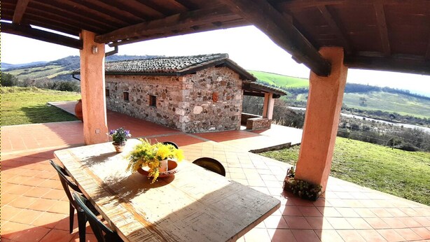 Un edificio de piedra rústico con tejado de baldosas, un porche cubierto con una mesa y sillas, y vistas a colinas ondulantes al fondo.