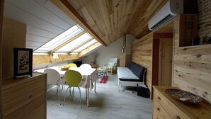 Una habitación acogedora en el ático, con techo inclinado, paredes de madera, tragaluces, una cama, una mesa con sillas y un pequeño sofá.