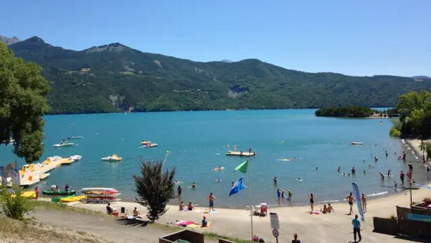 Una playa junto al lago con barcos, gente y montañas al fondo.