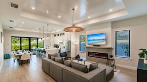 Una sala de estar amplia con un sofá seccional gris grande, una zona de comedor con mesa y sillas, una cocina con armarios blancos y una chimenea.
