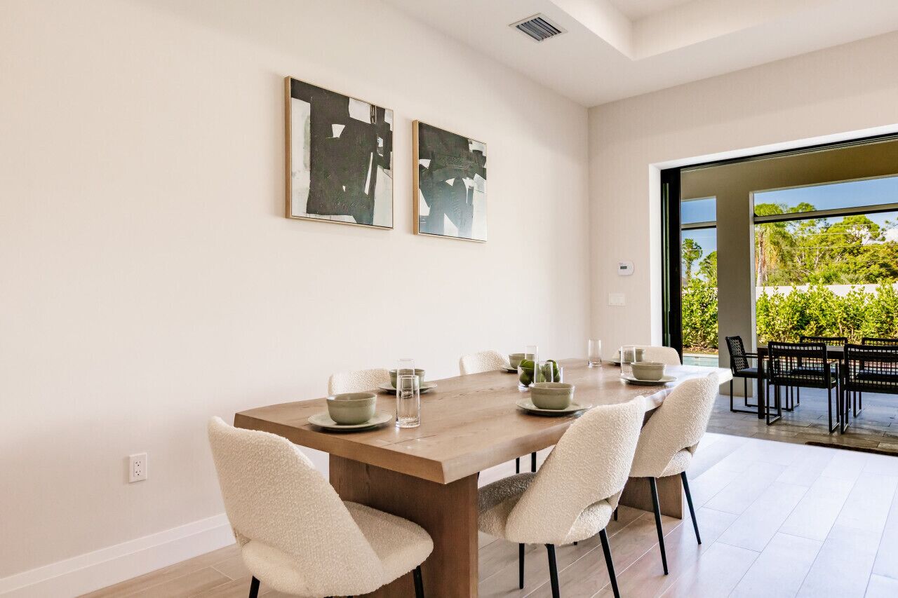 A modern dining area with a wooden table set for four, surrounded by chairs. There are two abstract paintings on the wall, and a view of greenery through a large window.