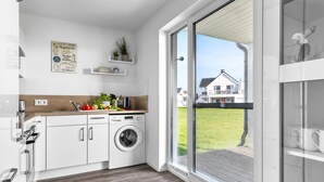 Cozinha moderna com máquina de lavar embutida, bancada de madeira e vista para uma casa por uma porta de vidro deslizante.