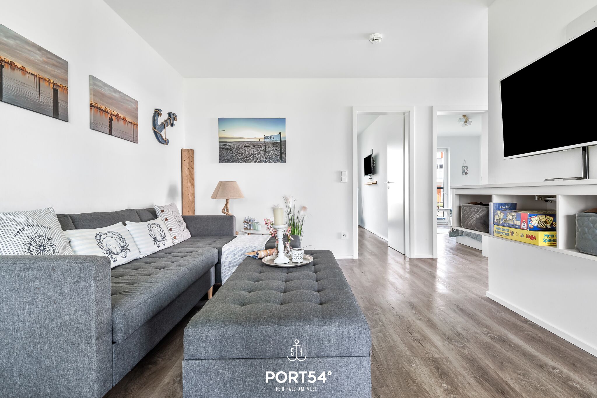 A modern living room with a grey sofa, a grey ottoman, and a flat-screen TV. There are wall-mounted artworks and a wooden accent piece.