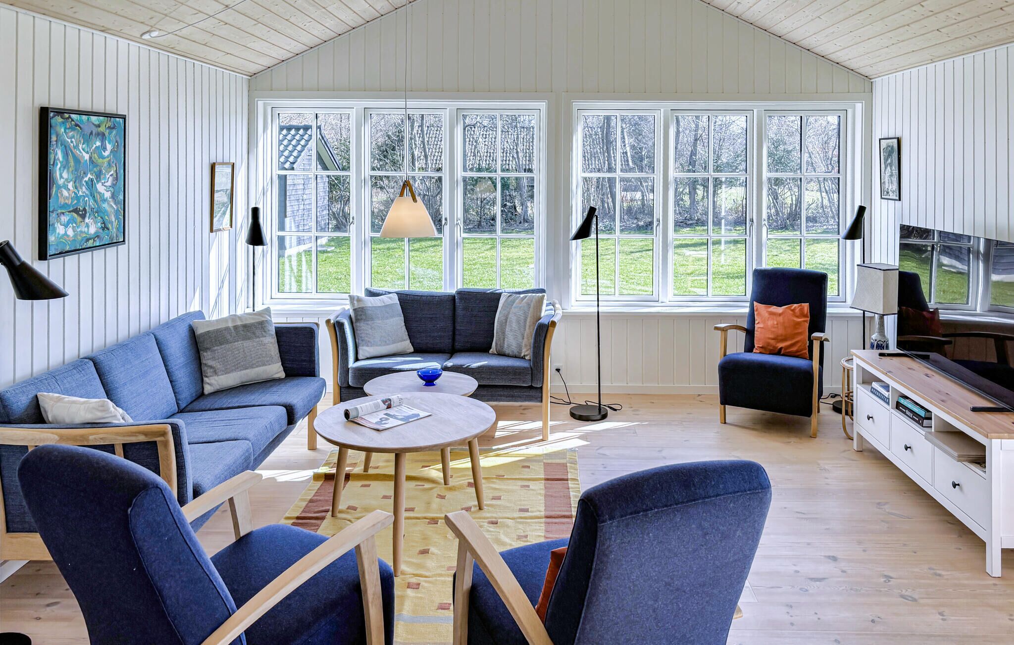 Ein geräumiges Wohnzimmer mit einer Couch, Sesseln, einem Couchtisch und einem Fernseher. Große Fenster bieten Blick ins Freie.