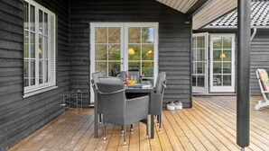 A wooden deck with two wicker chairs and a table, a window with a view of trees, and a door to the right. - Awesome home in Græsted with WiFi (Græsted)
