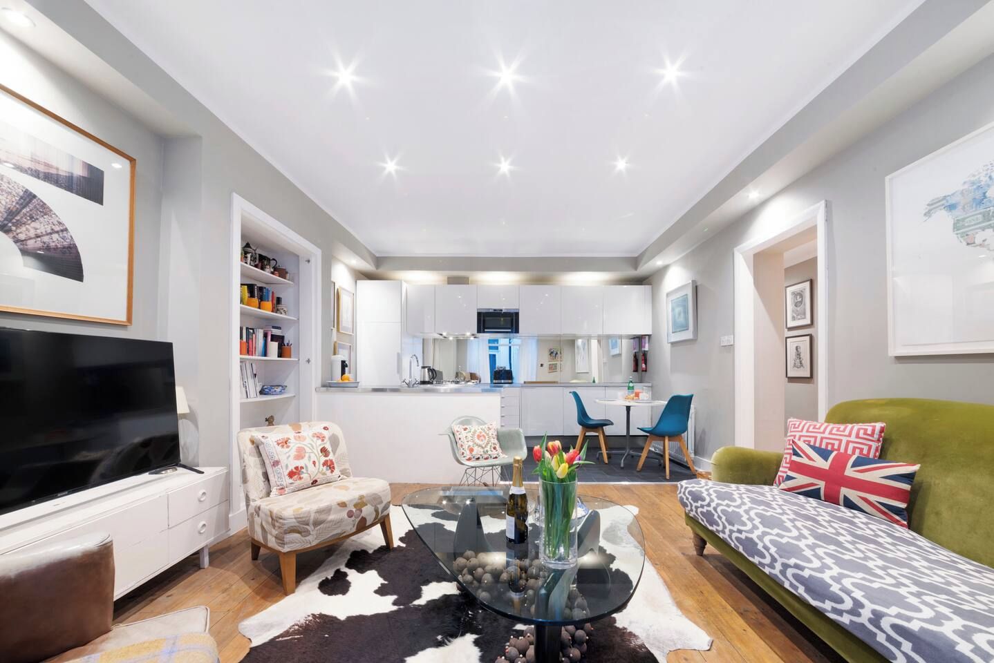A modern living room with a flat-screen TV, a sofa, a glass coffee table, and a dining area with chairs.