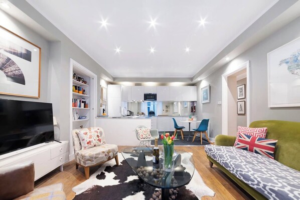 A modern living room with a flat-screen TV, a sofa, a glass coffee table, and a dining area with chairs.