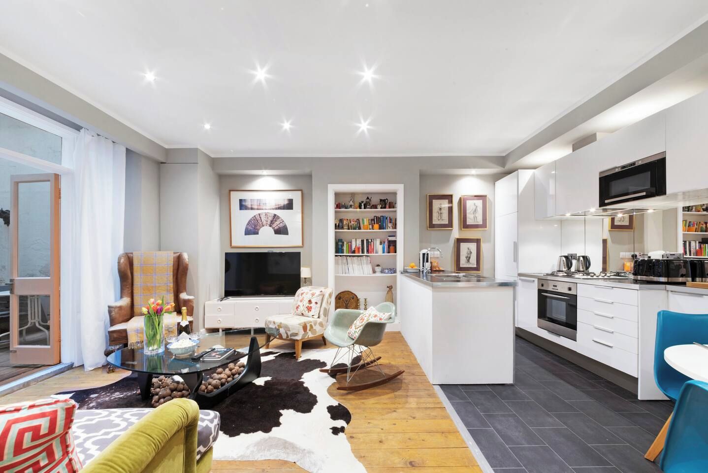 Uma sala de estar moderna com sofá, poltrona e mesa de centro, uma cozinha com eletrodomésticos e uma área de jantar, e uma grande janela.