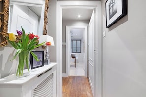A hallway leading to a bathroom with a white door, a framed picture on a console table, and a vase with flowers.