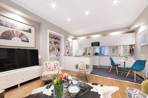 A modern living room with a flat-screen TV, a dining area with blue chairs, and a kitchen with white cabinets.