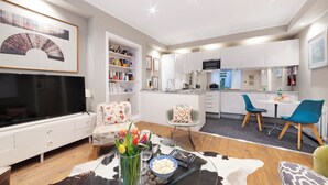 Ein modernes Wohnzimmer mit Flachbildfernseher, Couchtisch mit Dekoration, einer Couch mit floralen Kissen und einem Essbereich mit blauen Stühlen.